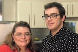 A woman in her mid-40s wears a red shirt and stands with her twenty-something yo son in their kitchen.