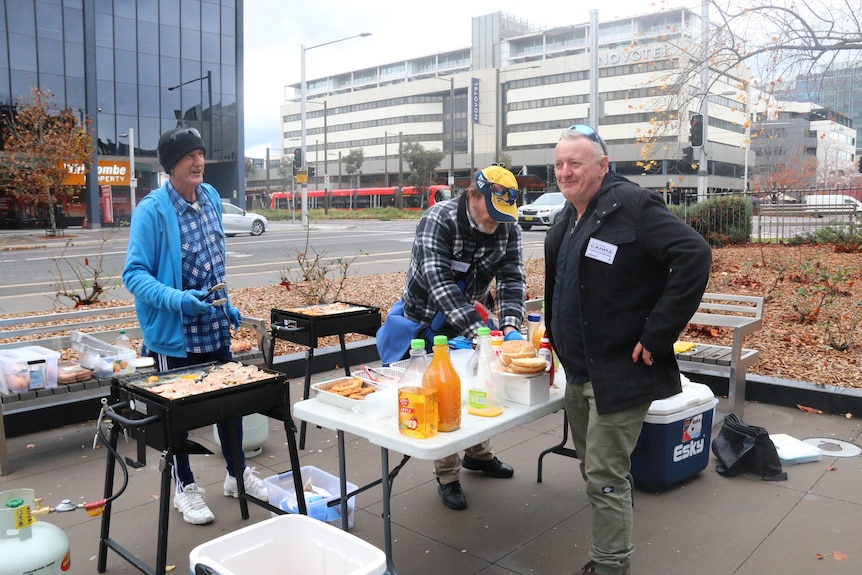 Three men stand at a table and barbecue and cook eggs and bacon in the Canberra city centre