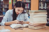 A young woman in a library looking at her smartphone, ignoring the pile of books in front of her