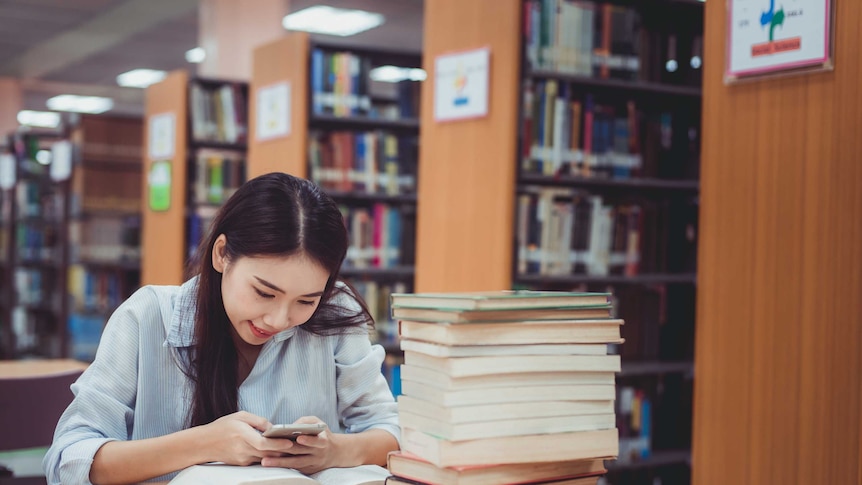 A young woman in a library looking at her smartphone, ignoring the pile of books in front of her