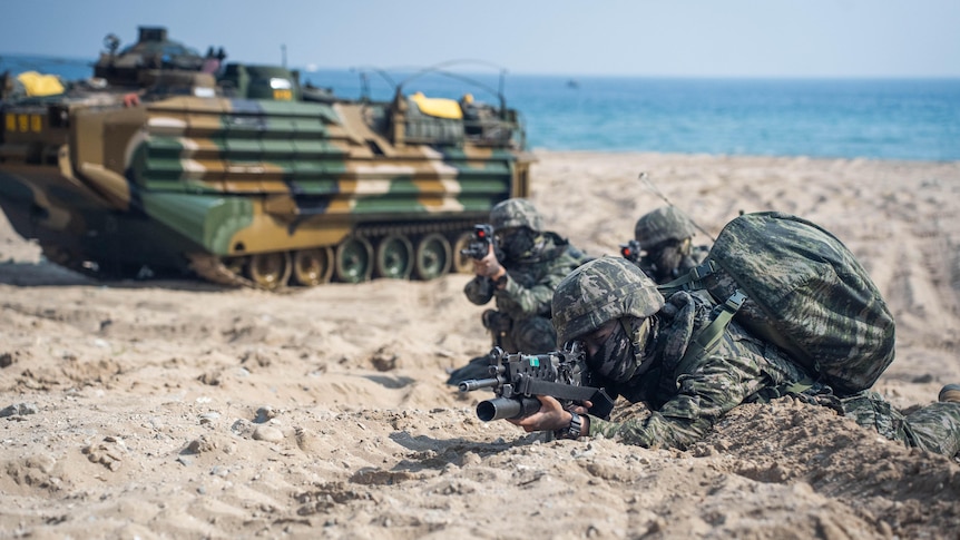 Three people in camouflage gear crouch on a beach in front of an amphibious assault vehicle
