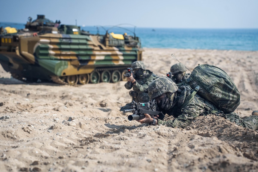 Three people in camouflage gear crouch on a beach in front of an amphibious assault vehicle