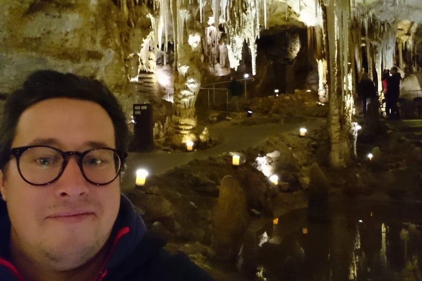 A man wearing glasses smiles at the camera with rock formations inside a cave behind him