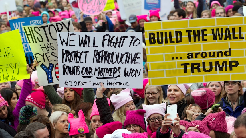 Women with bright pink hats and signs begin to gather for an anti-Trump march