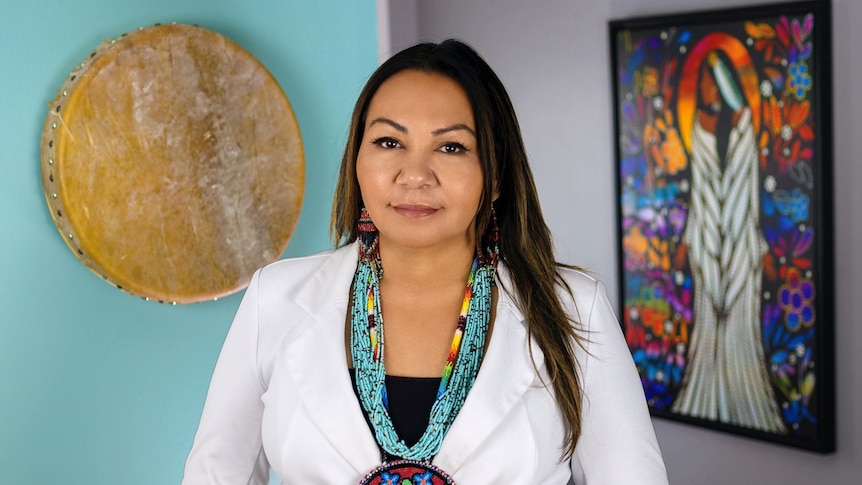 Sheila is wearing a white blazer and black top, She has a blue beaded necklace on. Behind her is a traditional artwork