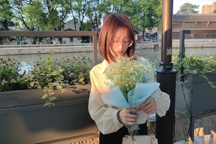 A young bespectacled Asian woman looks at a bunch of flowers she is holding.