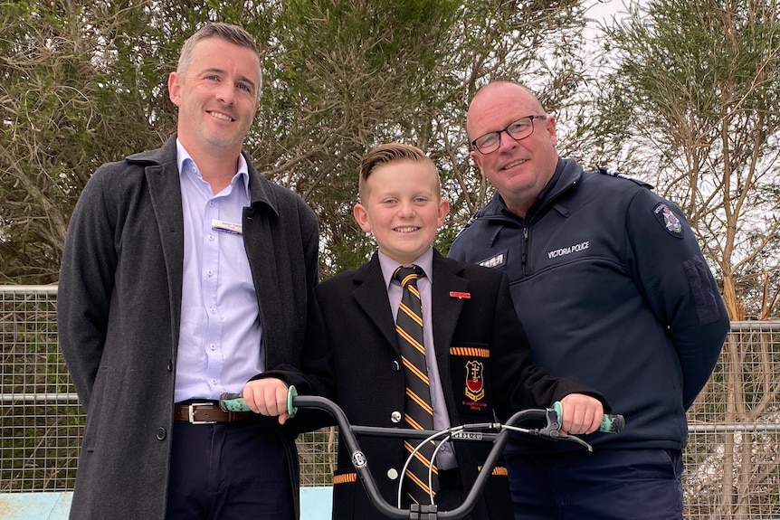 Boy on bike in school uniform flanked by two men in skate park.