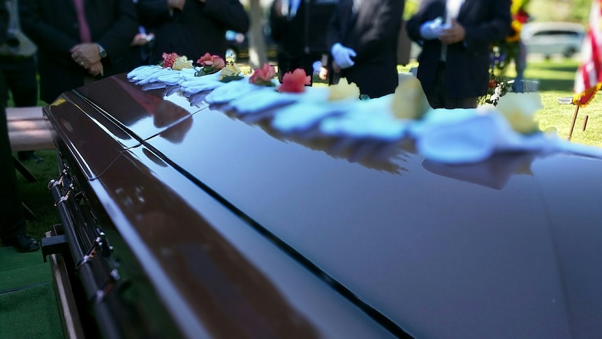 close up of coffin with row of flowers, people wearing suits in the background