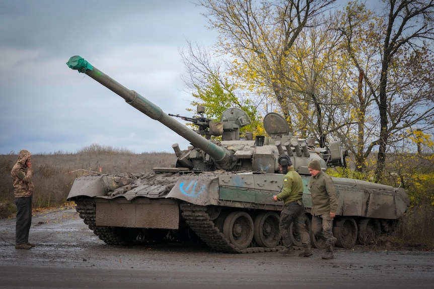 Three soldiers stand next to a tank on the side of the road.