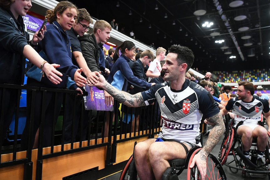 An English wheelchair rugby league player wheels along inside a stadium, with his hand out to high-five a group of young fans.