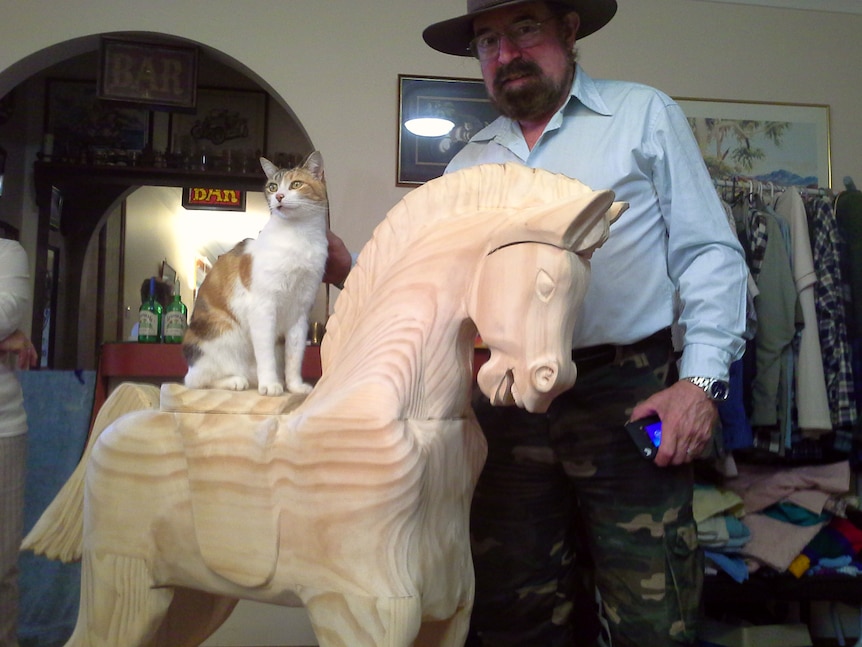Man stands behind unpainted rocking horse that has a white and ginger cat sitting on its back.