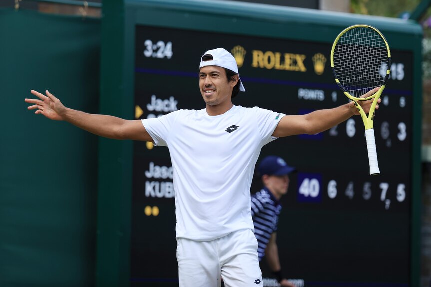 Australia's Jason Kubler stands on court with his arms wide in celebration at Wimbledon with the scoreboard behind him. 