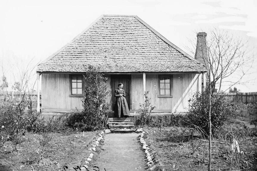 A black and white photo of a woman standing outside a colonial cottage.
