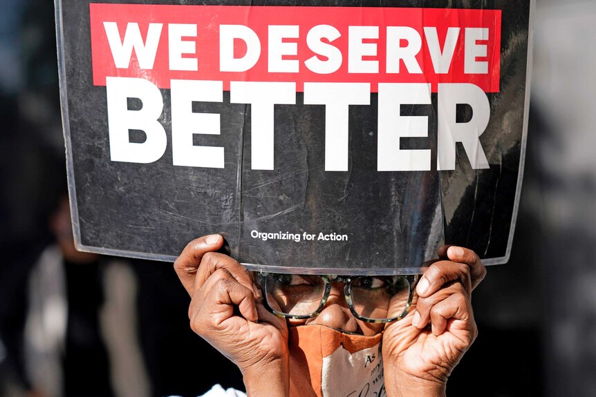 A woman wearing glasses and a mask holds up a sign saying We Deserve Better