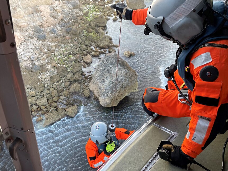 A helmeted paramedic in a brightly-coloured jump suit is winched down a cliff face.