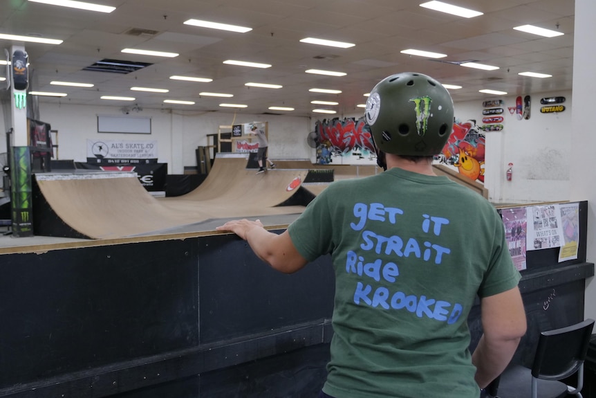 A woman in a green helmet and tshirt looks out at an indoor skatepark. Her shirt reads "get it strait, ride krooked"