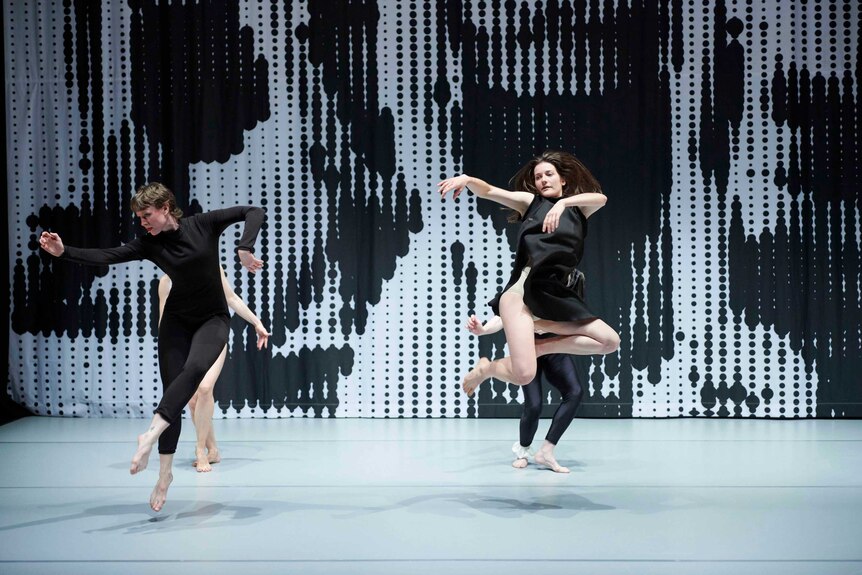 Four dancers on stage in black outfits, the front two leap in the air, black and white backdrop
