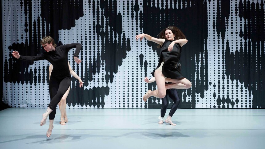 Four dancers on stage in black outfits, the front two leap in the air, black and white backdrop