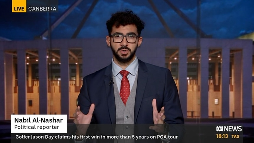 Bearded man wearing glasses, tie, suit and waistcoat doing a TV cross with Parliament House facade in background.