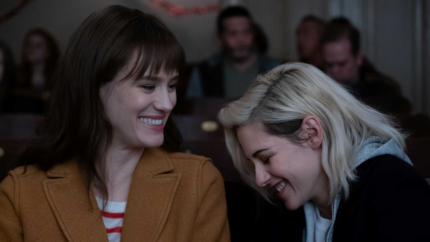 Actors Mackenzie Davis and Kristen Stewart sitting in a movie cinema laughing in the movie Happiest Season