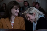 Actors Mackenzie Davis and Kristen Stewart sitting in a movie cinema laughing in the movie Happiest Season