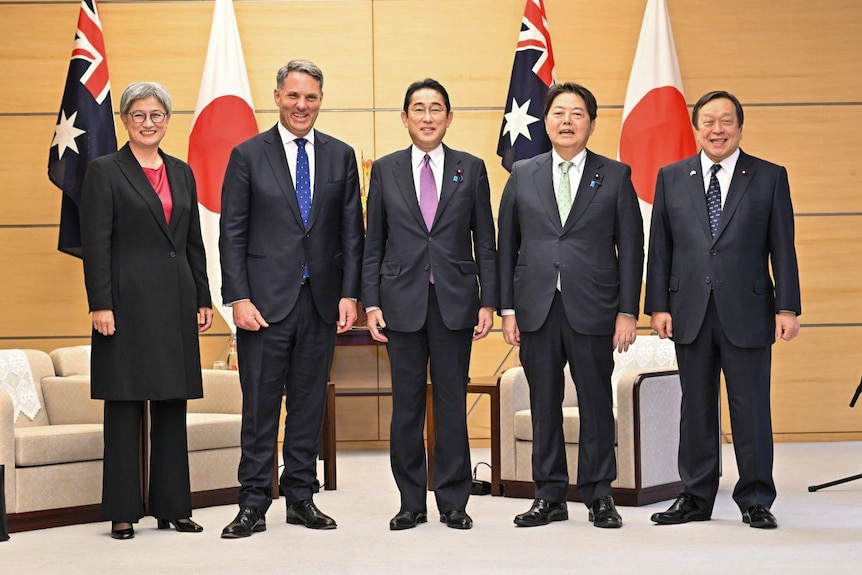 A woman on the left and four men on the right pose for photos in front of Australian and Japanese flags.
