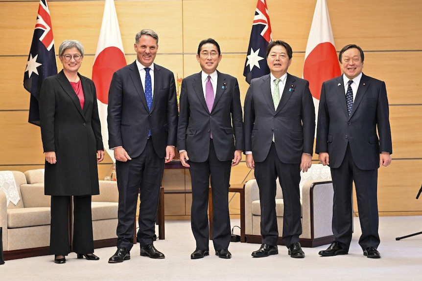 Woman on left and four men on right pose for photographs in front of Australian and Japanese flags.