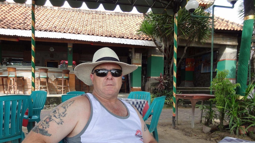 A man sitting at some plastic chairs at an outdoor bar. He wears a Bintang singlet and straw fedora.