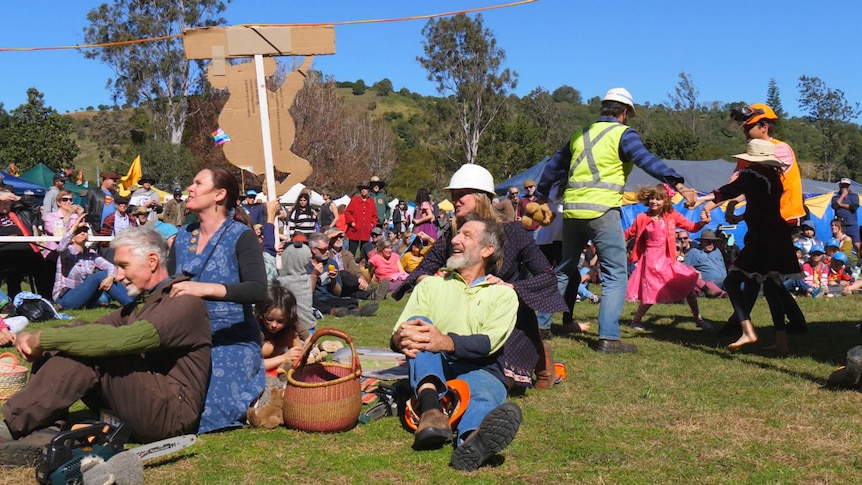 Two women sitting on grass massaging shoulders of workmen and kids spinning in a circle surrounded by crowds