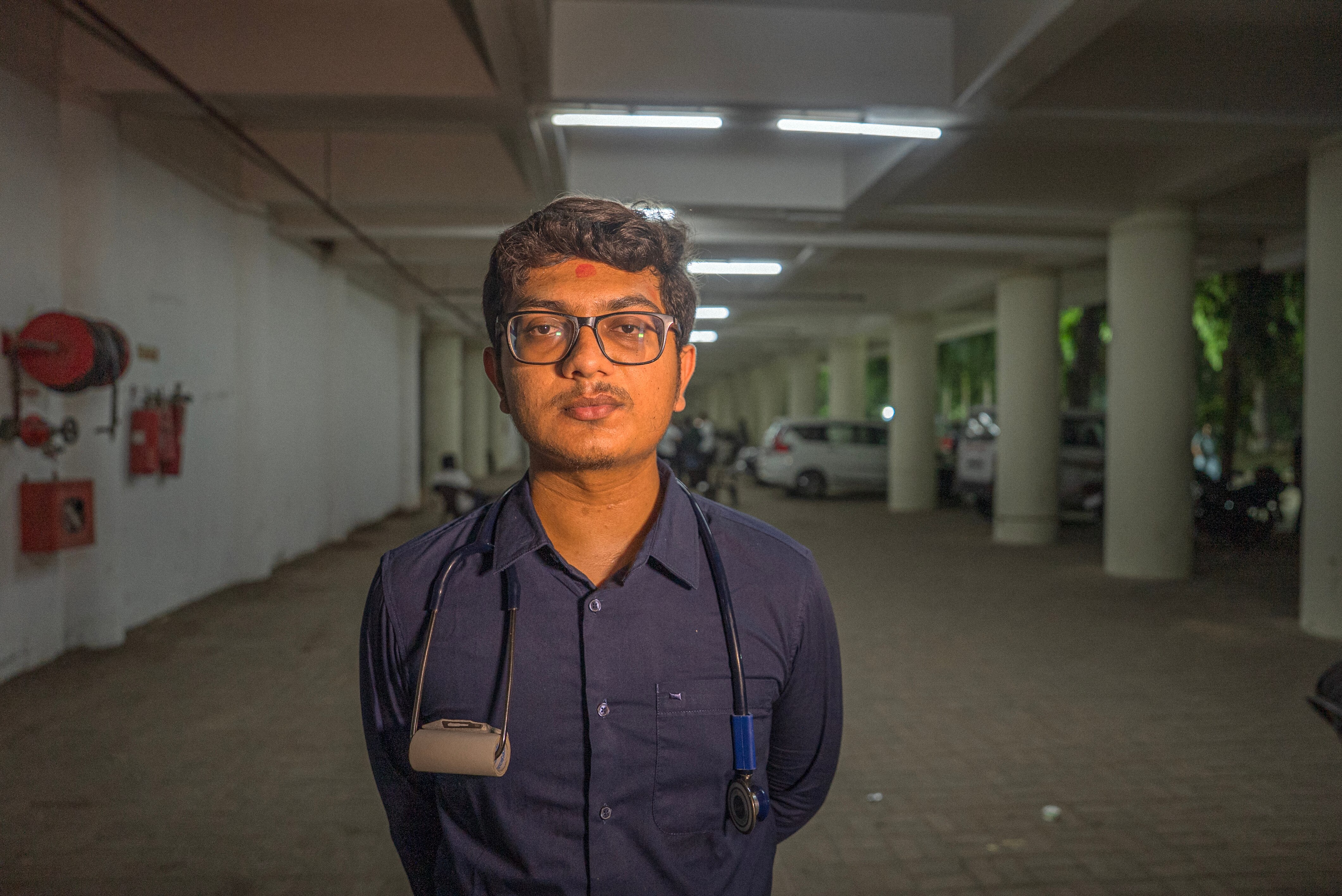 Young Indian man in a navy shirt with a stethoscope around his neck, standing in a carpark.
