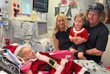 A small blonde child in a hospital bed with two adults. 