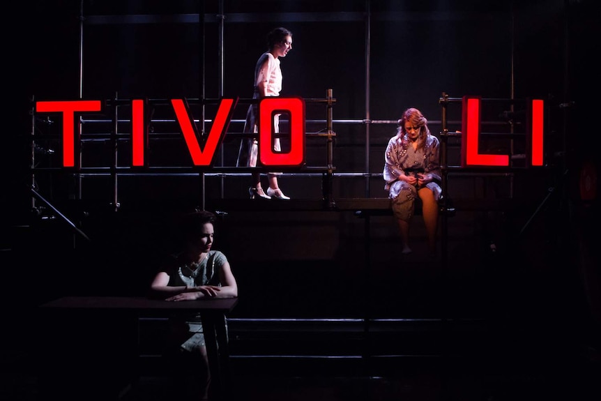 Two women sit on scaffolding with large red light-up letters spelling TIVOLI on it, while a third sits on stage below.