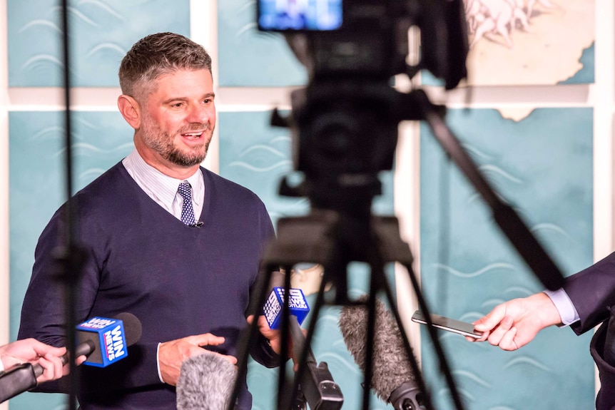 Nick Mitzevich at a press conference with microphones pointed at him.