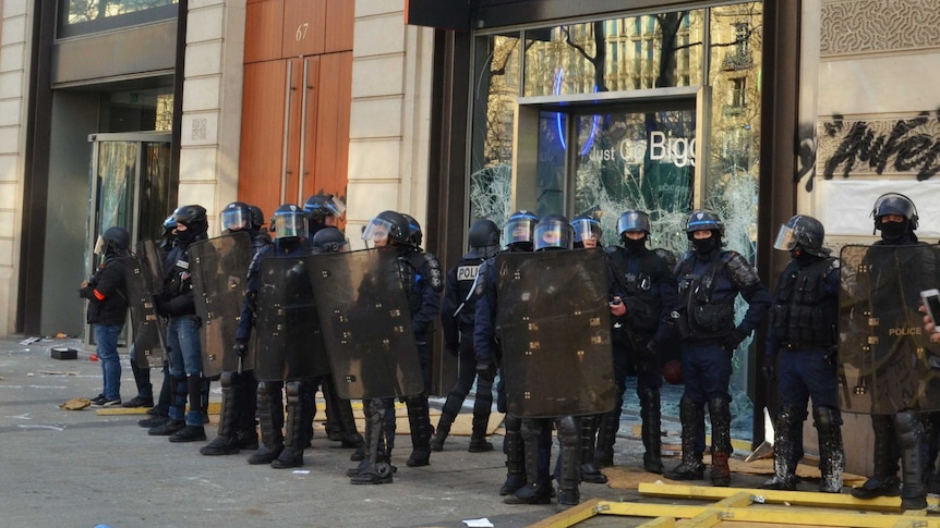 A group of police dressed in riot gear stand outside a smashed store window.