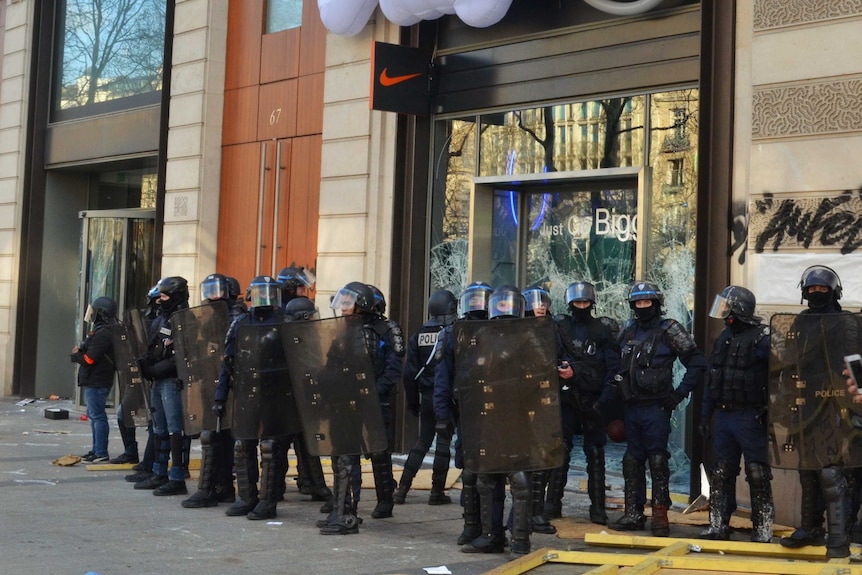 A group of police dressed in riot gear stand outside a smashed store window.