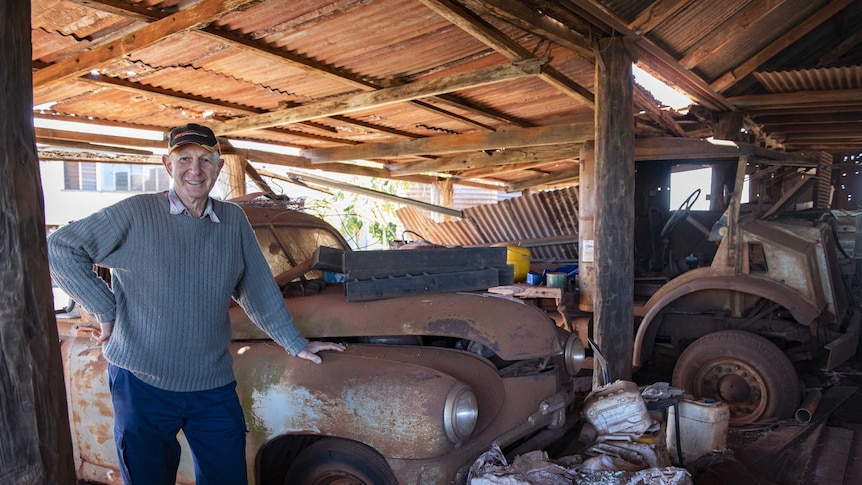 Mid shot of older man stands with his hand on the wheel hub of a rusted antique car in an old timber shed