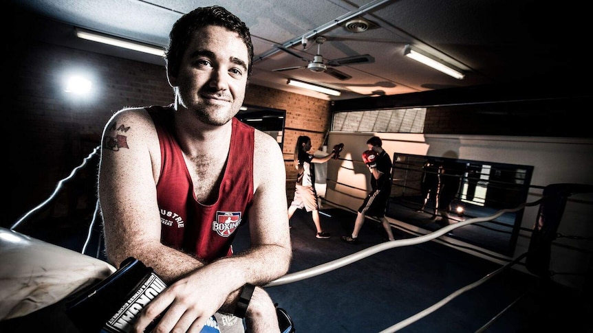 Kevin Rourke stands holding boxing gloves leaning on a boxing ring.