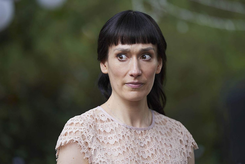 A woman with a blunt fringe and a deeply uncomfortable look on her face stands in front of a blurred green background.