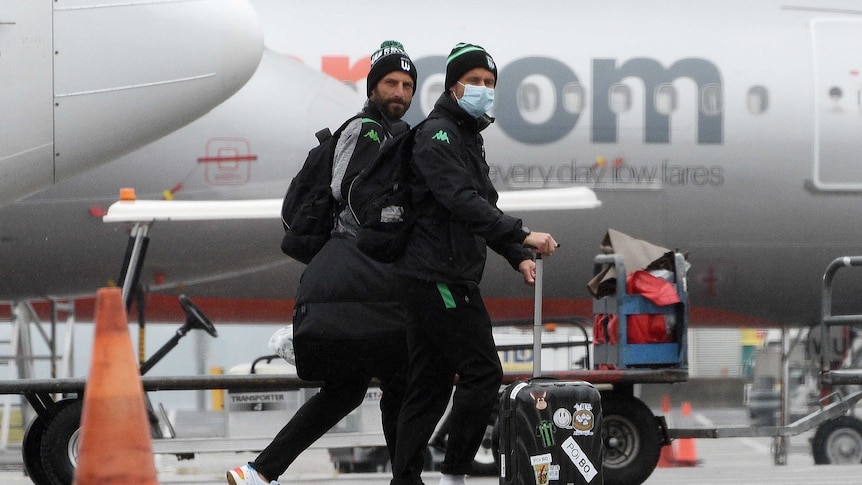 Two Western United A-League players arrive at Sydney Airport and walk across the tarmac with their luggage, wearing masks.