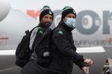 Two Western United A-League players arrive at Sydney Airport and walk across the tarmac with their luggage, wearing masks.