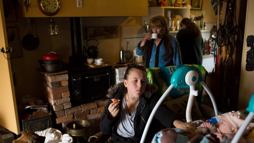 A woman makes breakfast as a younger woman can been seen with a baby in the background in a brick kitchen