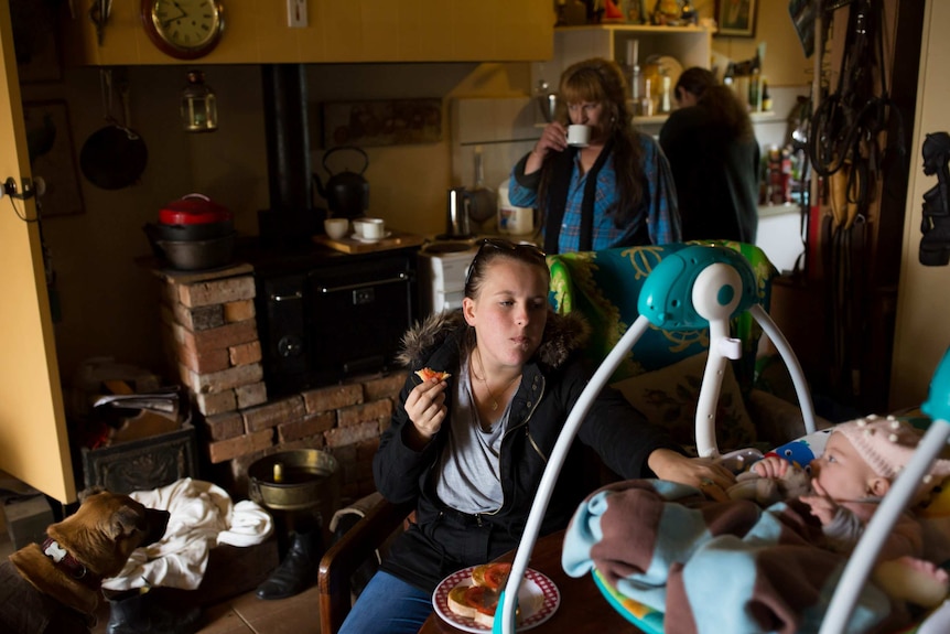 A woman makes breakfast as a younger woman can been seen with a baby in the background in a brick kitchen