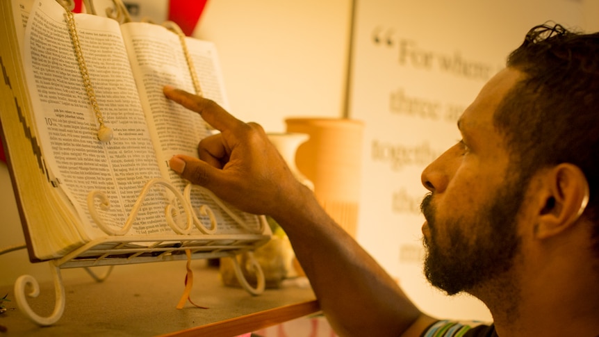 An Indigenous man studies the pages of a bible in a book holder on top of a bookcase.