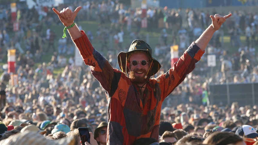 A man in a huge music festival crowd throws his arms akimbo, a big smile on his face.
