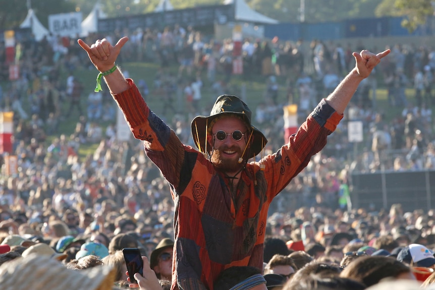 A man in a huge music festival crowd throws his arms akimbo, a big smile on his face.