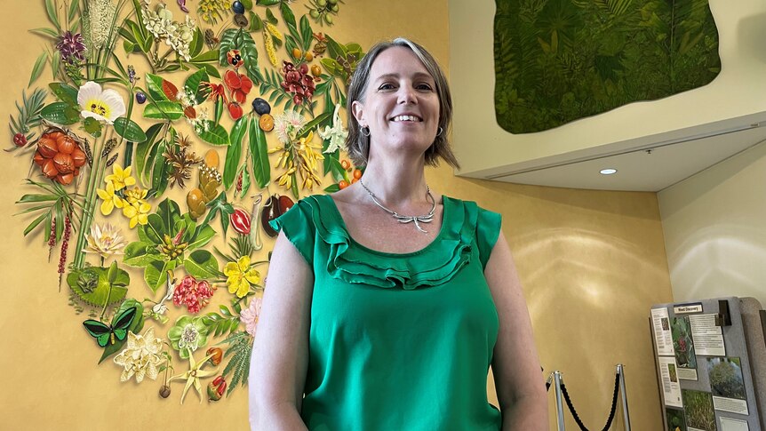 A woman with short blonde hair wears a green shirt with a ruffle and stands in front of a large floral mural.