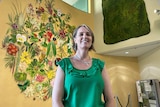 A woman with short blonde hair wears a green shirt with a ruffle and stands in front of a large floral mural.