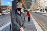 A woman with short blonde hair wearing a black mask waiting at a tram platform