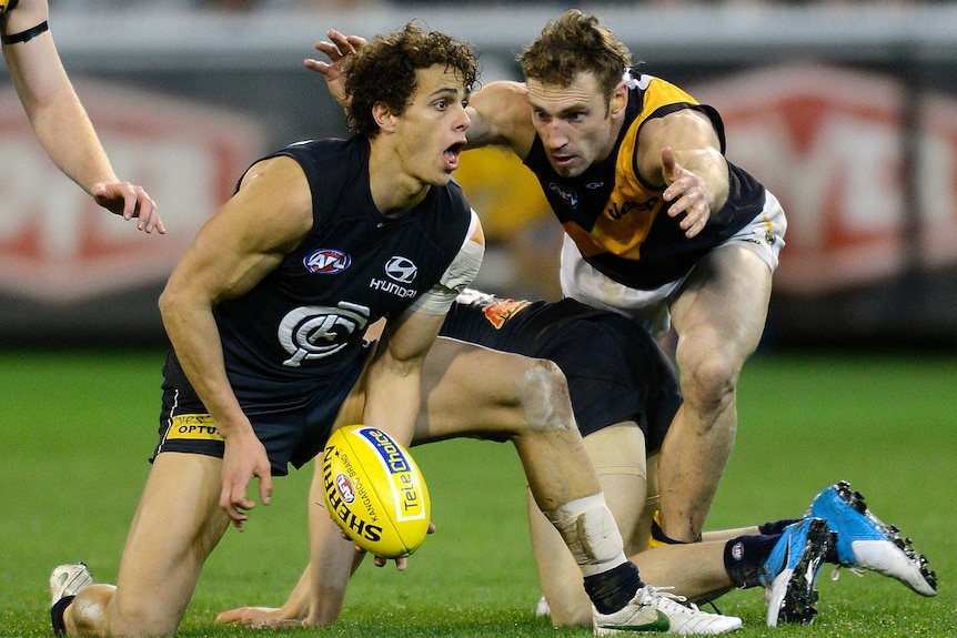 A Carlton AFL player attempts to handball while kneeling and under pressure from a Richmond opponent at the MCG.