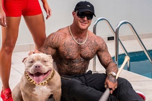 A heavily tattooed man holding a baseball bat sits next to a dog and a pool.